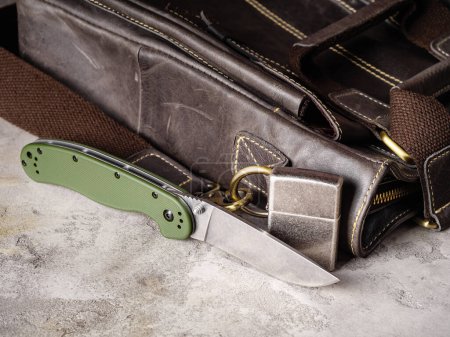 Foto de Green folding EDC knife and scratched lighter near a leather casual bag - Imagen libre de derechos