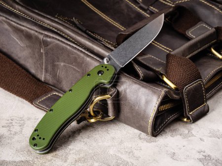 Foto de Green folding EDC knife with stonewash coated blade near a leather casual bag - Imagen libre de derechos