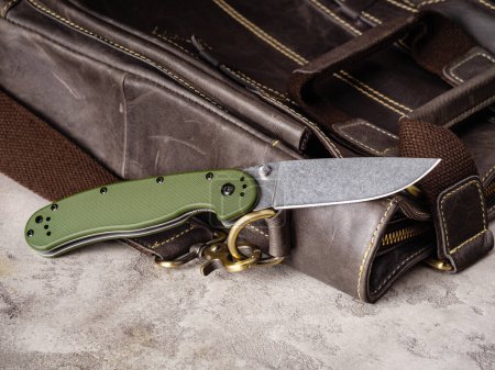 Foto de Green folding EDC knife near a vintage leather casual bag - Imagen libre de derechos