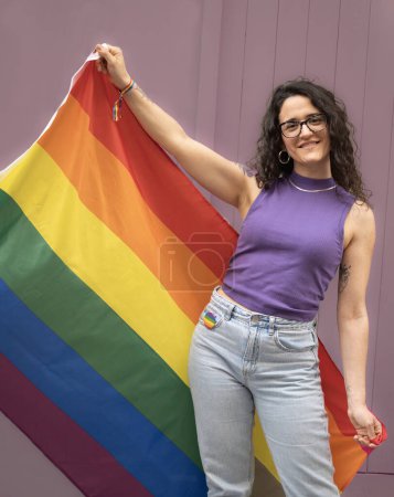 Téléchargez les photos : Vue de face portrait d'une jeune femme lesbienne heureuse tenant un drapeau arc-en-ciel sur le côté. Concept de fierté LGBT. - en image libre de droit