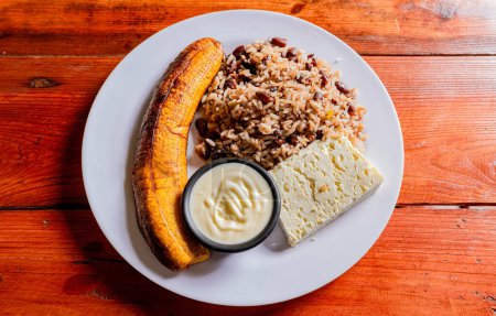 Photo for Gallopinto plate with cheese and maduro on wooden table. Nicaraguan food concept, Traditional Gallo Pinto meal with maduro and cheese served - Royalty Free Image