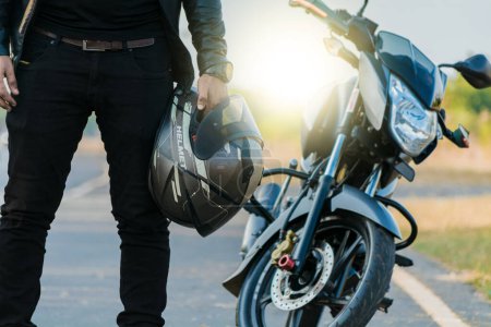 Photo for Unrecognizable biker holding helmet next to motorbike near the road. Motorcyclist holding safety helmet next to his motorbike near the road. Motorcycle safety helmet concept - Royalty Free Image
