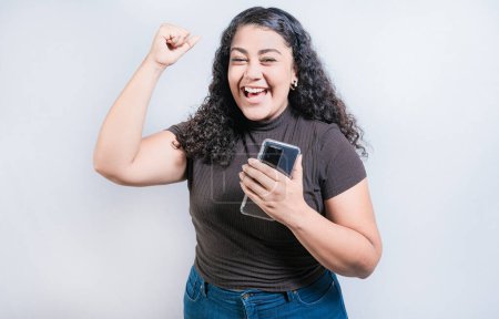 Foto de Ganar feliz joven mujer sosteniendo el teléfono celular. Feliz chica latina celebrando con teléfono aislado. Gente feliz sosteniendo smartphone y celebrando - Imagen libre de derechos