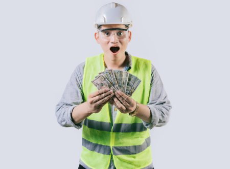 Ingeniero sorprendido en casco sosteniendo dinero aislado. Ingeniero varón sorprendido sosteniendo billetes sobre fondo aislado