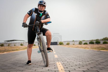 Cycliste souriant sur son vélo regardant la caméra sur la route. Chubby cycliste masculin en vêtements de sport chevauchant un vélo à l'extérieur