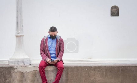 Tipo con máscara quirúrgica sentado con su teléfono celular, hombre con máscara sentado fuera con su teléfono celular