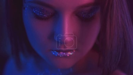 Téléchargez les photos : Neon light face. Party makeup. Nightclub beauty. Closeup portrait of purple blue color glow woman with closed eyes shimmering eyeshadow lips on dark. - en image libre de droit
