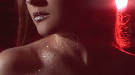 Téléchargez les photos : Glitter makeup. Glamour look. Disco beauty. Cropped closeup of woman face with shimmering shiny skin metallic lips in neon red color night light on dark. - en image libre de droit