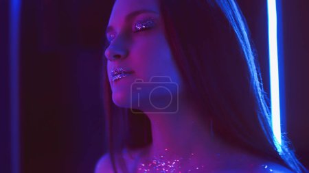 Foto de Neon girl. Glitter makeup. Nightclub fashion. Purple blue color glow relaxed woman with sparkling face skin eyeshadow lips in blur fluorescent light on dark free space. - Imagen libre de derechos