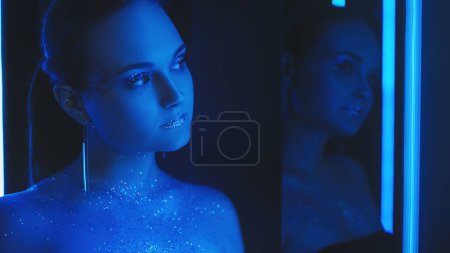 Photo for Neon girl. Sparkling makeup. Futuristic beauty. Night portrait of blue color light woman with shimmering glitter face skin eyeshadow lips in mirror on dark. - Royalty Free Image