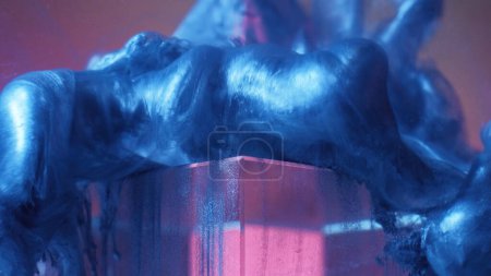 Foto de Paint drop on glass cube. Ink water. Smoke cloud. Defocused blue color shiny fluid splash on pink transparent angle abstract background. - Imagen libre de derechos