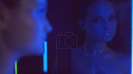Photo for Disco makeup. Neon portrait. Nightclub beauty. Blue purple color light woman with sparkling glitter face skin metallic lips in mirror on dark. - Royalty Free Image