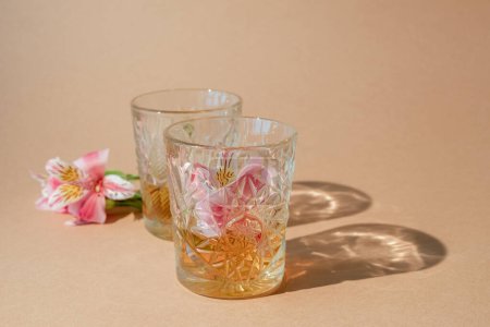Photo for Summer drink. Alcoholic beverage. Herbal liquor. Couple of brown whiskey glasses with flower decor on beige copy space background. - Royalty Free Image