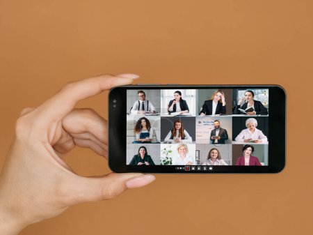 Photo for Video meeting. Mobile briefing. Distant communication. Business team working on phone screen in female hand on brown copy space background. - Royalty Free Image