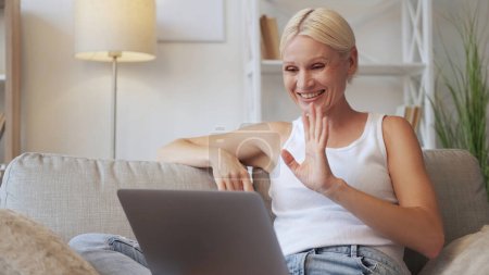 Photo for Video call. Online chat. Distance communication. Relaxed happy cheerful mature woman waving hello using laptop on cozy couch at modern home interior with free space. - Royalty Free Image