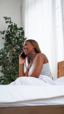Photo for Morning call. Mobile communication. Happy amused smiling woman enjoying fun conversation talking on phone in bed in light home bedroom interior. - Royalty Free Image