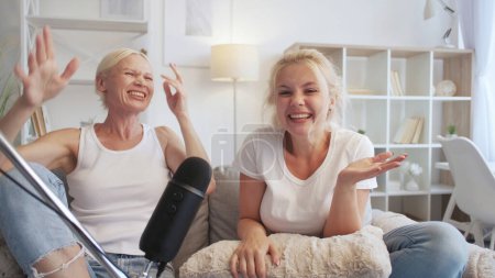 Fun conversation. Interview podcast. Family communication. Two relaxed women laughing recording audio into microphone on couch at home studio.