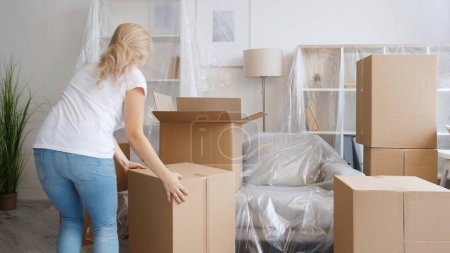 Home relocation. Moving day. Removal service. Unrecognizable woman carrying carton boxes in light modern apartment with covered furniture.