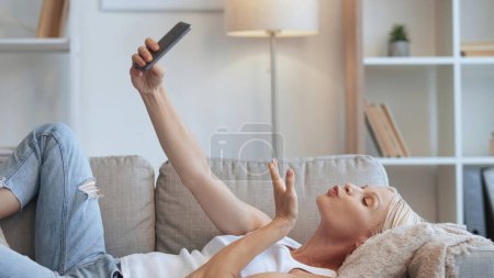 Photo for Romantic chat. Video call. Online flirt. Distance relationship. Relaxed mature woman blowing air kiss on phone selfie camera lying on sofa in modern home interior with free space. - Royalty Free Image