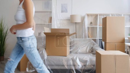 Photo for Home moving. Flat relocation. Removal service. Unrecognizable woman leaving room with carton boxes in modern apartment with polyethylene covered furniture. - Royalty Free Image