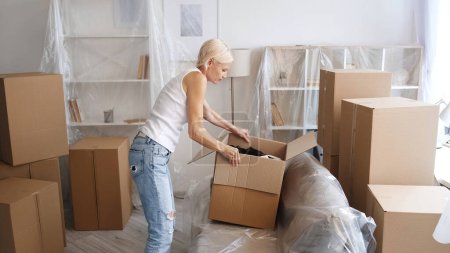 Photo for Relocation stress. Moving belongings. Tired senior woman carrying unpacking things from carton boxes in light modern home with covered furniture. - Royalty Free Image