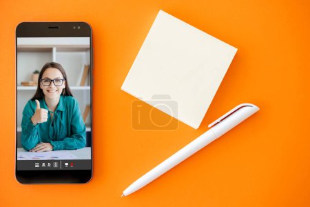 Photo for Online expert. Video coaching. Mobile conference. Satisfied business woman approving with thumb up like gesture on phone screen on orange copy space background with pen empty note. - Royalty Free Image