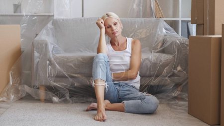 Photo for Sad relocation. Moving home. Eviction stress. Upset depressed tired pensive mature woman thinking of relocation in flat with boxes polyethylene covered furniture. - Royalty Free Image