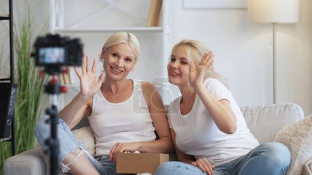 Photo for Female vlog. Product unboxing. Online streaming. Two cheerful smiling influencer women waving on camera on tripod recording video content on couch at home. - Royalty Free Image