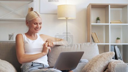 Téléchargez les photos : Week-end Gadget. Loisirs en ligne Divertissement Internet. Femme âgée souriante détendue appréciant regarder un film en utilisant un ordinateur portable sur un canapé dans le salon moderne avec espace libre. - en image libre de droit