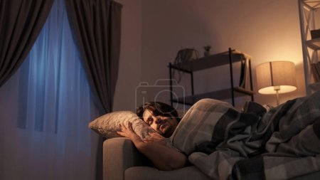 Foto de Noche de descanso. Relajación. Acogedora noche. Hombre tranquilo cansado durmiendo en almohada suave debajo de la manta en el sofá en el interior de la sala de estar oscura. - Imagen libre de derechos