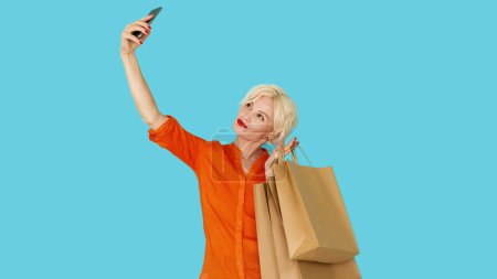 Photo for Mobile photo. Shopaholic lifestyle. Happy woman. Pretty smiling middle-aged lady shooting selfie on smartphone holding paper bags with purchases posing blue. - Royalty Free Image