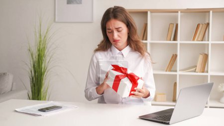 Undesirable gift. Upset woman. Corporate greeting. Gloomy disappointed lady holding present box red ribbon sitting desk in light room interior.