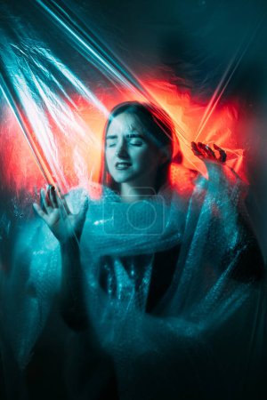 Photo for Plastic pollution. Global warming. Ecology problem. Red blue color light defocused woman suffocating in bubble wrap trapped in wrinkled polyethylene film. - Royalty Free Image