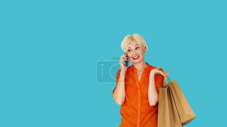 Téléchargez les photos : Communication mobile. Femme inspirée. Rabais shopping. Happy lady parler smartphone tenant des sacs en papier avec des achats sur l'espace de copie bleue. - en image libre de droit