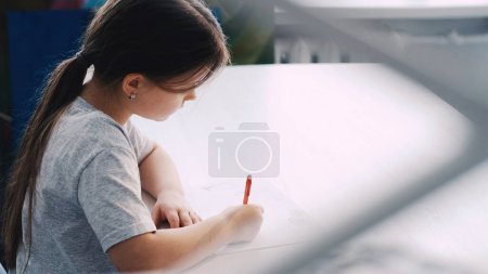 Téléchargez les photos : Studio d'art. Petite fille. Cours de peinture. Dessin d'enfant inspiré avec crayon sur papier blanc à l'intérieur de la pièce lumineuse floue. - en image libre de droit