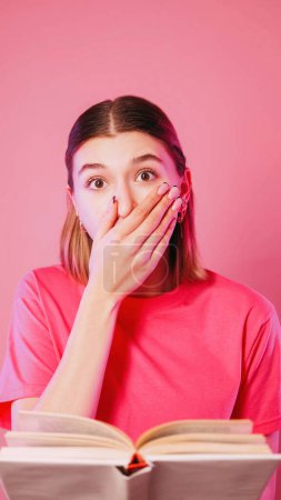 Photo for Surprising book. Astonishing reading. Shocked girl covering mouth amazed by novel end look with frightened eyes isolated on neon color pink background. - Royalty Free Image