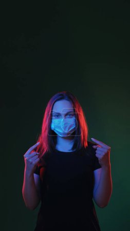 Photo for Covid protection. Coronavirus hygiene. Flu epidemic. Red blue color light girl pointing at protective mask on face on dark green empty space background - Royalty Free Image