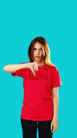 Photo for Dislike gesture. Displeased woman. Dissatisfied girl in red T-shirt showing thumb down expressing bad choice isolated on cyan blue copy space background. - Royalty Free Image