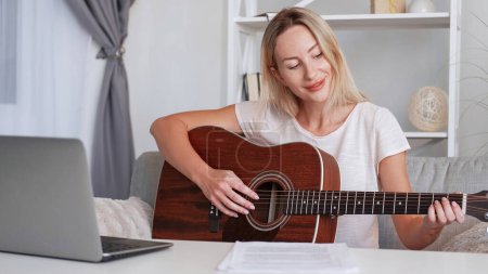 Photo for Online guitar tutorial. Music lesson. Diligent studious woman learning chords playing instrument watching video on laptop home interior. - Royalty Free Image