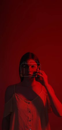 Photo for Phone conversation. Concerned woman portrait. Confused concentrated neon color light girl mobile conversation isolated on dark red copy space background. - Royalty Free Image