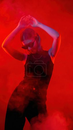 Photo for Red smoke portrait. Night dancing girl. Satisfied stylish woman in black sunglasses raised arms enjoying disco time on neon light background. - Royalty Free Image