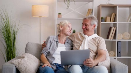 Photo for Family leisure. Happy couple. Online movie. Excited middle-aged man and woman sitting sofa with laptop in light home interior. - Royalty Free Image