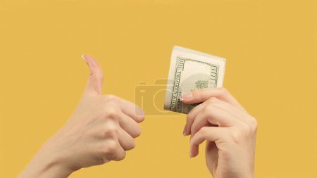 Téléchargez les photos : Revenus passifs. Un geste d'accord. Mains de femme montrant des dollars et comme signe d sur fond jaune orange. - en image libre de droit