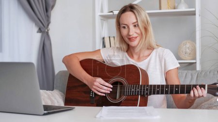 Photo for Online guitar class. Music tutor. Smiling pleased woman teacher tutorial lesson teaching playing instrument recording educational video on laptop camera home interior. - Royalty Free Image