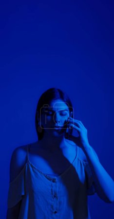 Photo for Mobile talking. Serious woman portrait. Focused neon color light girl smartphone call attentive expression listening information isolated on dark blue empty space background. - Royalty Free Image