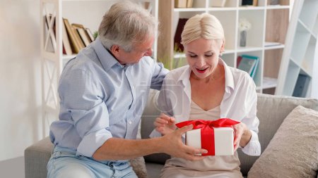 Photo for Happy anniversary. Amazed woman. Holiday surprise. Festive middle-aged wife getting pleasant unexpected gift box from husband in light home interior. - Royalty Free Image