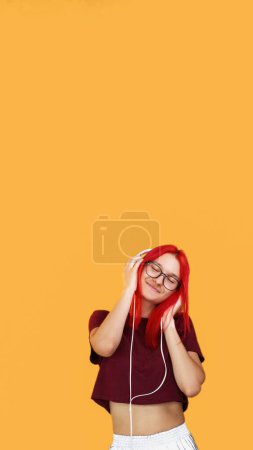 Foto de Escucha la canción. Música de ocio. Joven chica pelirroja sonriente complacida con auriculares de alambre blanco disfrutando del sonido en el fondo naranja espacio vacío. - Imagen libre de derechos