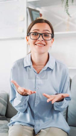 Photo for Video call. Discussion online. Internet communication. Young emotional smiling gesticulating smart woman in glasses and blue shirt in light interior at home. - Royalty Free Image