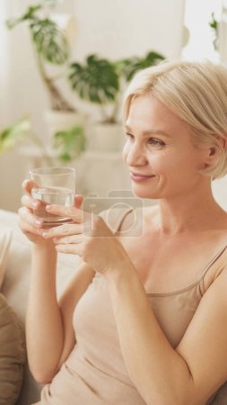 Photo for Healthy lifestyle. Happy woman. Morning treatment. Satisfied middle-aged lady enjoying glass of clear water sitting sofa light home interior. - Royalty Free Image