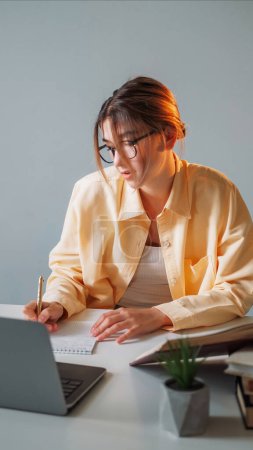 Photo for Female student. Distance education. Homework doing. Smart woman writing in copybook sitting desk opened laptop in light room interior. - Royalty Free Image